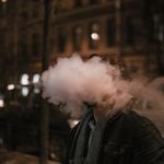 A man exhales a thick cloud of vapor on a city street at night, creating an artistic and dramatic effect.