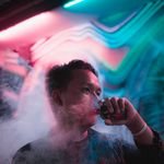 Asian man vaping in a colorful and smoky nightclub with vibrant neon lights.