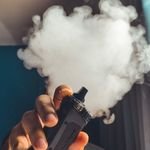 Hand holding a vaping device with a large cloud of smoke, indoors.