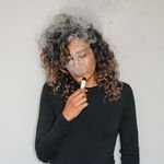 Portrait of a woman with curly hair vaping indoors, featuring smoke and a relaxed expression.