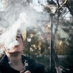 Portrait of a young man vaping in a park, releasing clouds of vapor on a clear day.